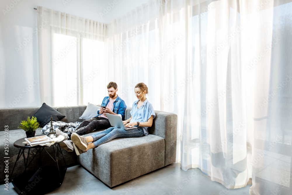Man and woman dressed casually in blue shirts sitting on the couch using laptop and phone at home. Wide view with copy space Man and woman dressed casually in blue shirts sitting on the couch using laptop and phone at home. Wide view with copy space