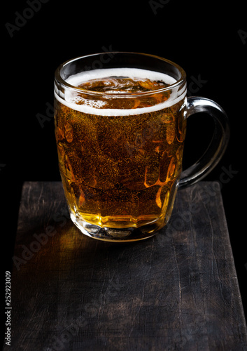 Glass of lager beer with foam and bubbles on vintage wooden board on black background.