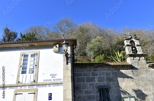 Ponte Maceira, Coruña, Spain. Wall with sundial, iron light and Camino de Santiago symbol. San Blas chapel bell tower with trees and blue sky. photo