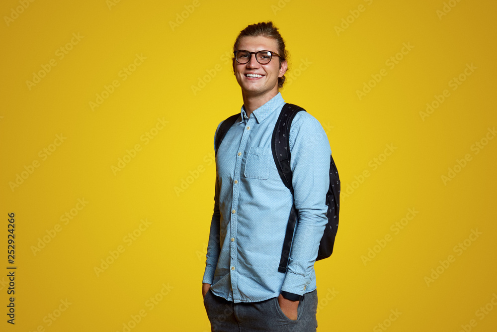 Portrait of a joyful young attractive student with backpack, wearing blue casual shirt and eyeglasses. Man standing and holding hands in pockets, laughing joyful over bright yellow background. Portrait of a joyful young attractive student with backpack, wearing blue casual shirt and eyeglasses. Man standing and holding hands in pockets, laughing joyful over bright yellow background.