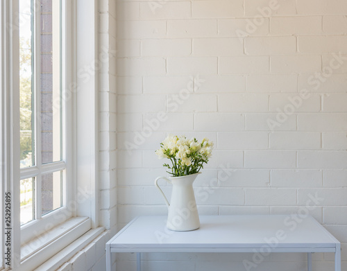 White freesias in jug on table against painted brick wall next to window with copy space