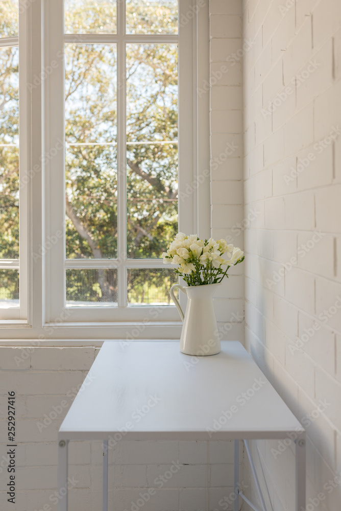 Vertical view of white freesias in jug on table against window and painted brick wall (selective focus) Vertical view of white freesias in jug on table against window and painted brick wall (selective focus)