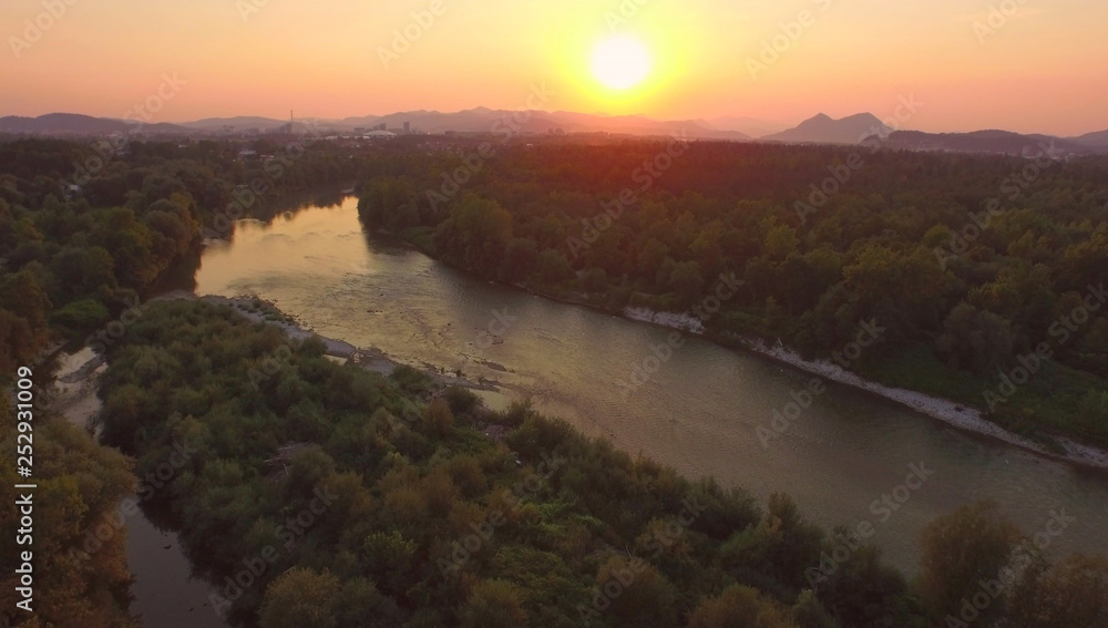 AERIAL: Flying over a calm river running through the green forest at sunset.