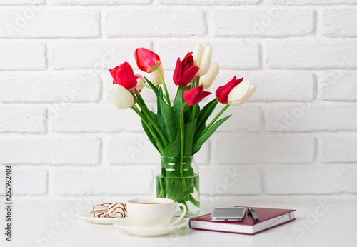 Fototapeta Naklejka Na Ścianę i Meble -  tulip flowers are in a vase on the table, cup of coffee, diary and smartphone, white brick wall as background