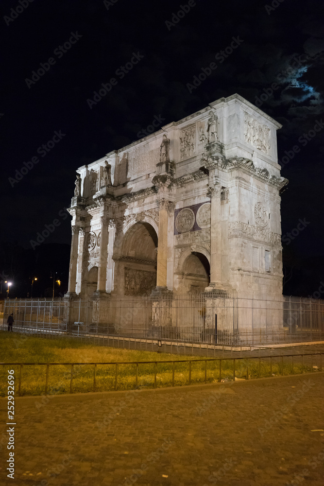 arch of constantine of the roman forum viewed through the gated arch of the passage at the entrance of the Roman Colosseum at night.
