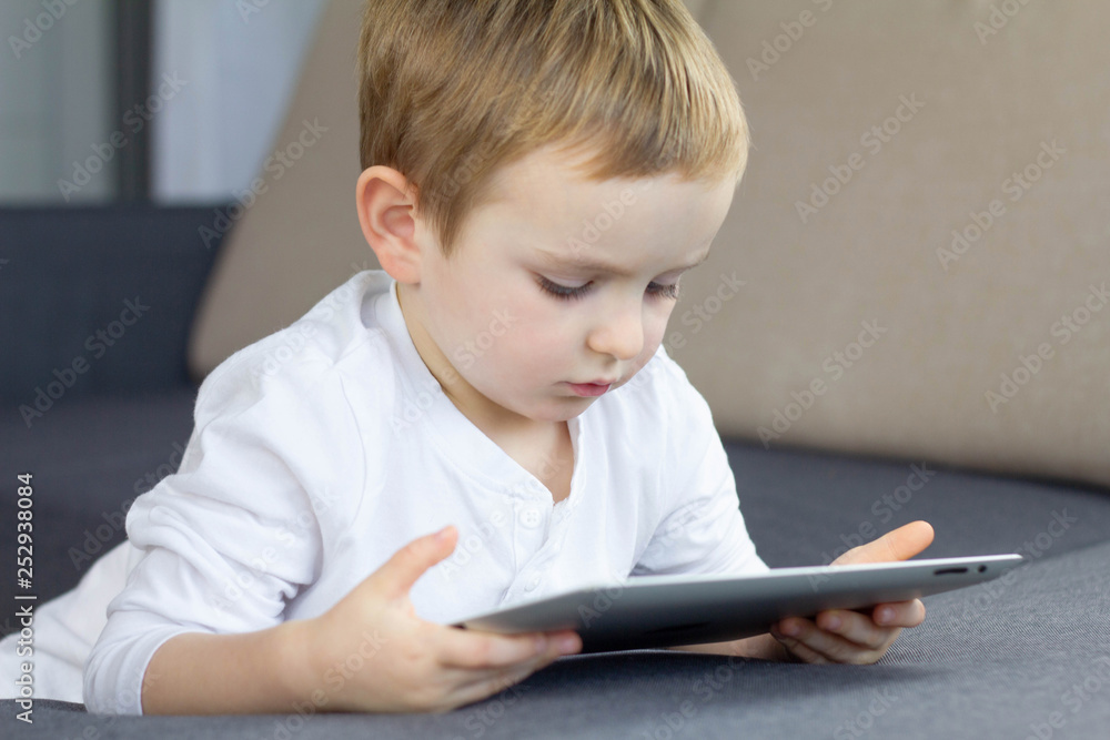 Little blonde boy using touchscreen tablet at home. Happy smart child watching tutorials or playing game on tablet computer. Education, technology, children's security and fun concept. Little blonde boy using touchscreen tablet at home. Happy smart child watching tutorials or playing game on tablet computer. Education, technology, children's security and fun concept.