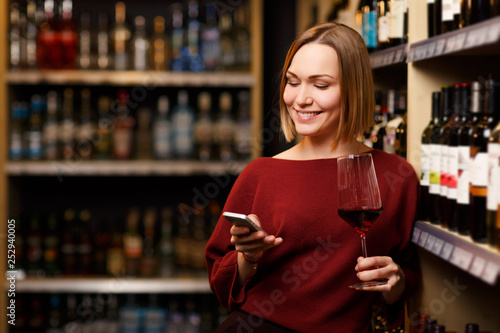 Image of happy blonde with phone and glass in hands at store with wine