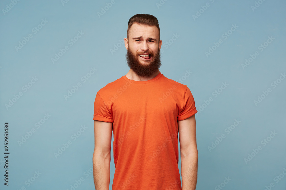 Portrait of handsome bearded young man with stylish haircut wears red t-shirt looking with disgust aversion and shows tongue, isolated over blue background Portrait of handsome bearded young man with stylish haircut wears red t-shirt looking with disgust aversion and shows tongue, isolated over blue background