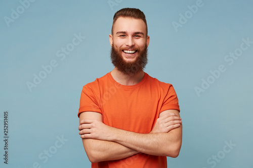 Studio portrait of emotional happy funny smiling boyfriend man with a heavy beard stands with arms crossed dressed in red t-shirt isolated over blue background