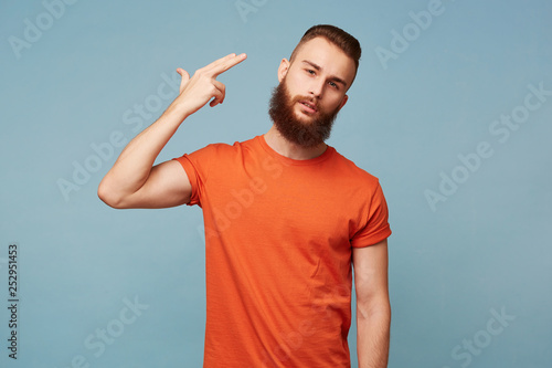 Portrait of young bearded man committing suicide with finger gun gesture. Portrait of despaired guy shooting himself making finger pistol sign against blue wall background. Human face expressions