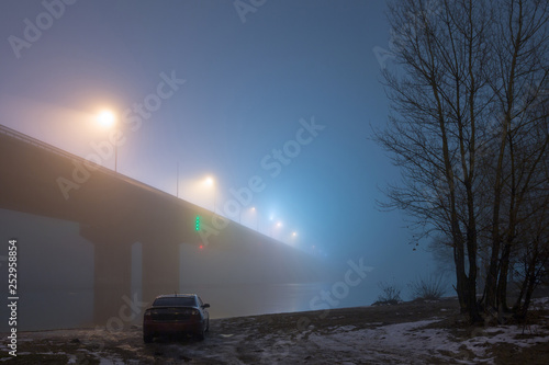 A mysterious evening fog above the river in big city. Bridge in the mist  cold weather scenery. Soft  blurry  misty look. Colorful  mystic industrial cityscape. A car on the river bank.