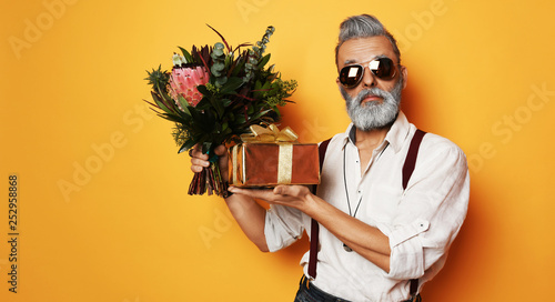 Brutal senior bearded old man in aviator sunglasses with a bunch of flowers and a gift