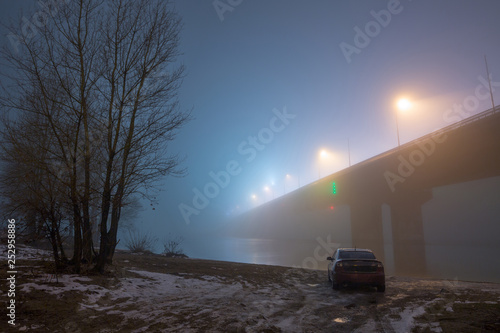 A mysterious evening fog above the river in big city. Bridge in the mist, cold weather scenery. Soft, blurry, misty look. Colorful, mystic industrial cityscape. A car on the river bank. © Sodel Vladyslav