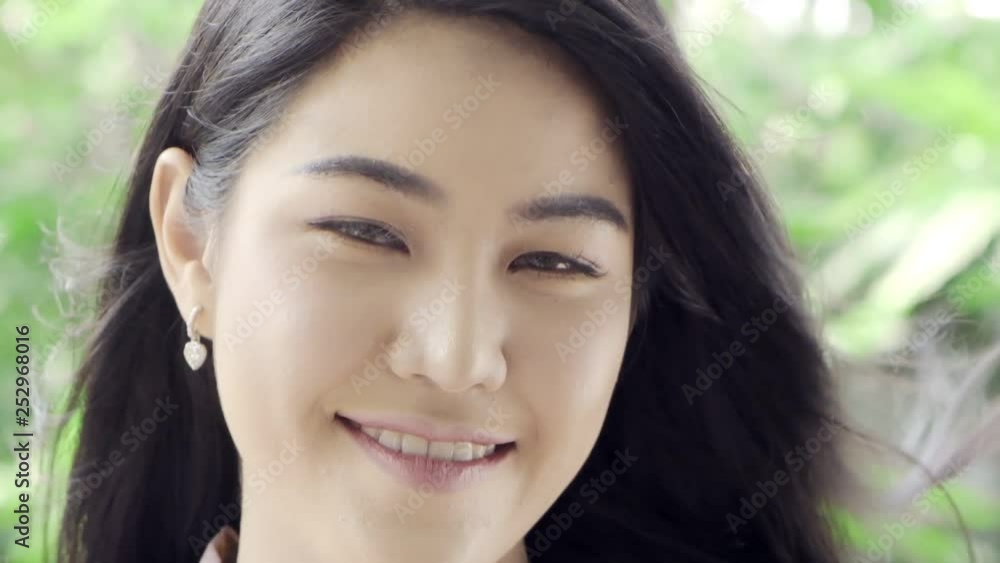 Super Slow motion of young beautiful Asian woman smile at camera with wind blow her hair in her home garden with green tree in background, close up shot. Millennial lifestyle concept. Super Slow motion of young beautiful Asian woman smile at camera with wind blow her hair in her home garden with green tree in background, close up shot. Millennial lifestyle concept.