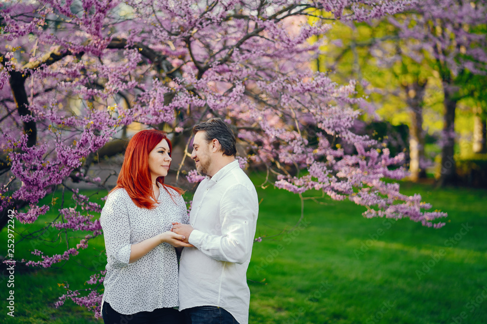 Fototapeta premium beautiful and stylish redhead woman in a white blouse standing in a sunny summer park with her handsome husband near flower trees