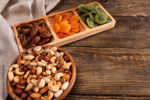 Dates, dried apricots and kiwis in a Compartmental dish and assortment of nuts in wooden bowl on a dark wooden table.