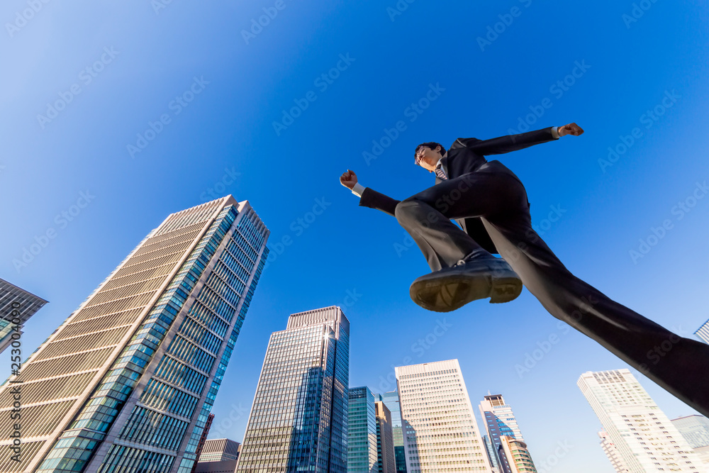 青空とオフィスビル群をバックにジャンプする若いビジネスマン1人。ビジネス、努力、成功、活発、エネルギッシュ、イメージ