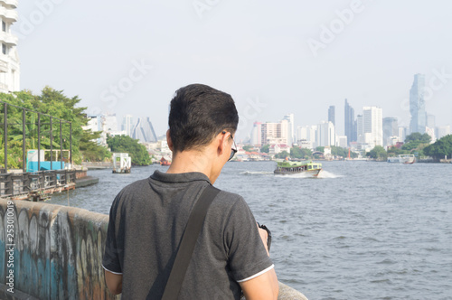 A man setting his camera before taking photos at the river with tall buildings and trees in distance
