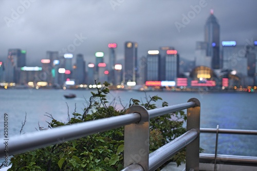 Metal handrail with blurred bokeh background of Hong Kong Island skyline cityscape and green leaves of bush under cloudy gray sky