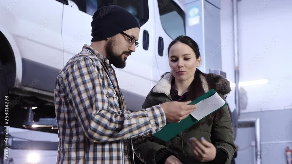 Auto service. A beautiful-looking girl, a brunette, signs documents with a car maintenance mechanic about receiving a car from repair Auto service. A beautiful-looking girl, a brunette, signs documents with a car maintenance mechanic about receiving a car from repair