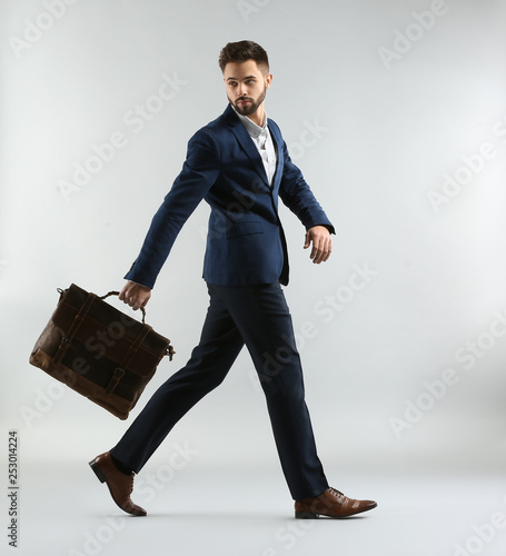 Fashionable young man in formal clothes and with bag walking against light ba...
