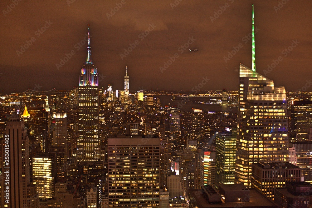 Fototapeta premium Illuminated Manhattan skyline at night time with skyscrapers, high-rise buildings, low-rise buildings, neon lights under gray cloudy sky with light pollution