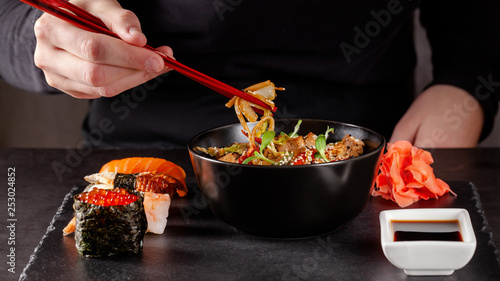 Concept of Asian cuisine. The girl is holding Japanese chopsticks in her hand and eating Chinese noodles from a black plate in a restaurant. background image. copy space