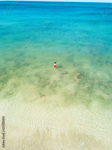 Aerial view of a girl in red swimming suit walking into the Andaman sea of green color. There is blue sky and white sand beach. Koh Lanta, Thailand, Asia. © dimabucci