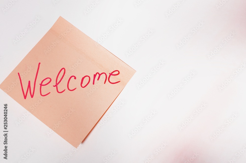 The sentence Welcome written by hand on a pink post it and put on the white table The sentence Welcome written by hand on a pink post it and put on the white table