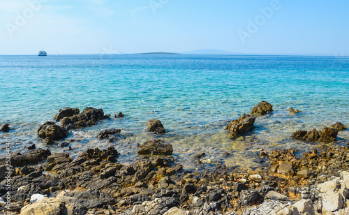 Beautiful blue and calm sea with small waves and boats on background on sunny day. Rocks and stones on the beach in island with hill and othe island on background.