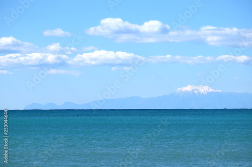 High mountains silhouettes with white snow cap on top far away on horizon in light haze after the sea under blue sky with white cumulus clouds