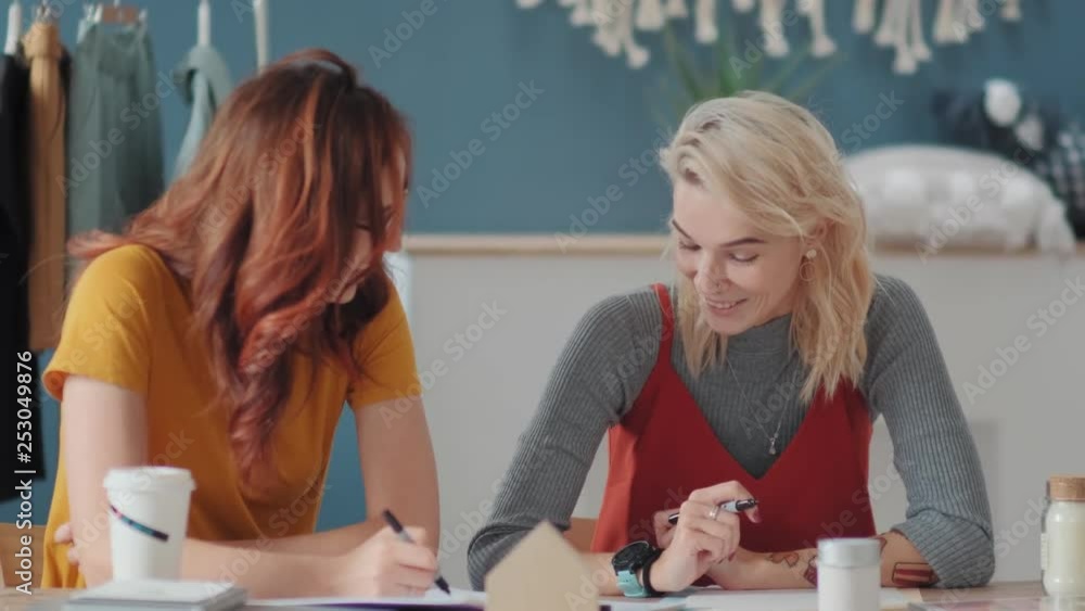 Two stylish young women discuss work in an informal, friendly atmosphere. Creative designers discuss the blog, new ideas for a collection of clothes. Work of two women at desk in a creative atmosphere