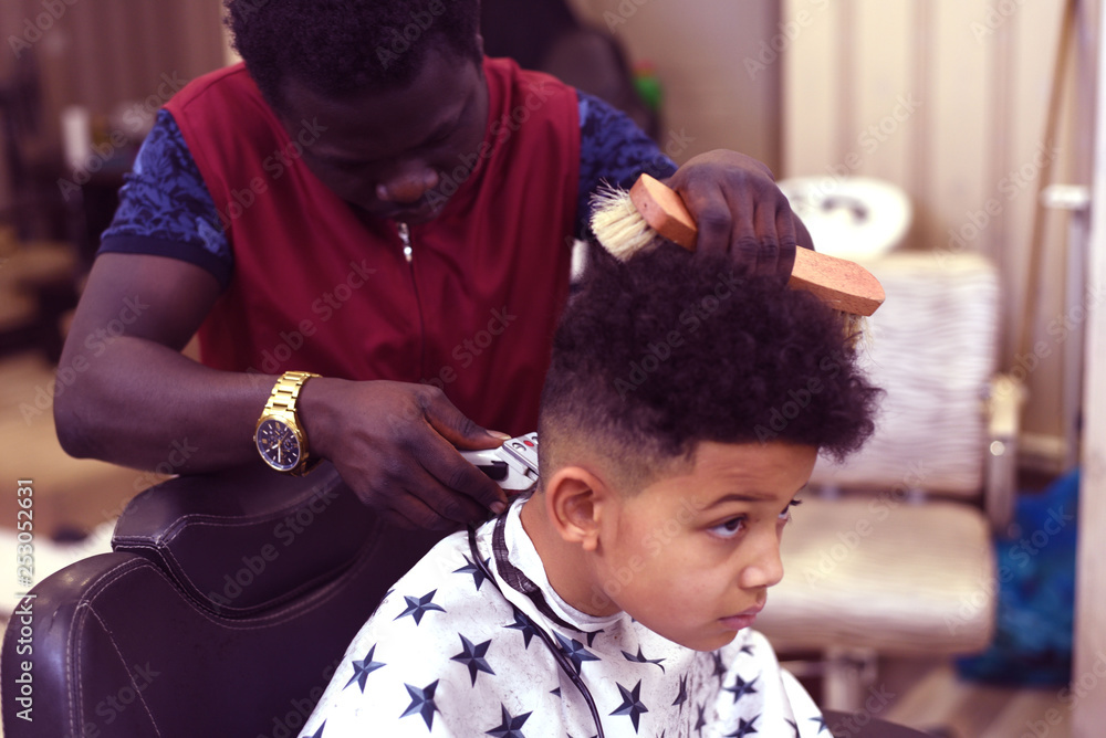Fotografia do Stock: Cute african american boy in the african barbershop.  Cute mixed boy makes a haircut in the African salon. Hair style. Haircut by  machine for children. School boy hair style.