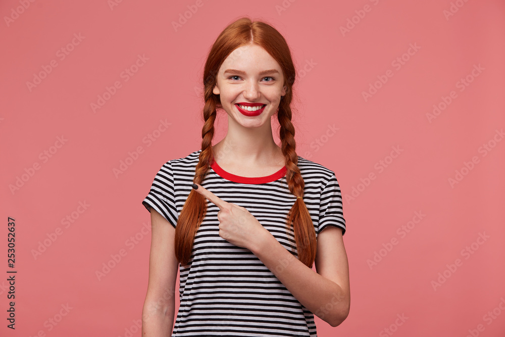 Close up of happy inspired red haired teen girl shows with index finger to the left side, is delighted with the product,advises to pay attention, shows place for your advertisement or promotional text Close up of happy inspired red haired teen girl shows with index finger to the left side, is delighted with the product,advises to pay attention, shows place for your advertisement or promotional text