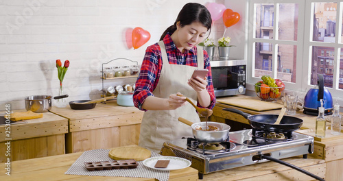 elegant girl stirring melted chocolate in liquid on stove hot water pot. young woman holding spoon and cellphone taking photo recording down process video of making dessert. handmade valentine gift