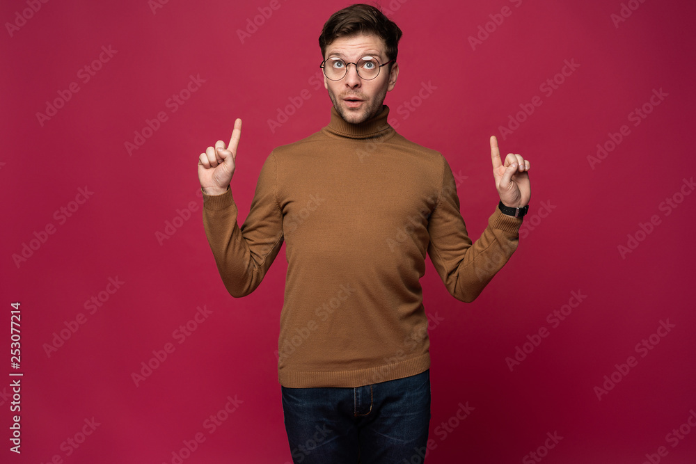 Portrait of a happy young man pointing fingers up at copy space isolated over pink background.
