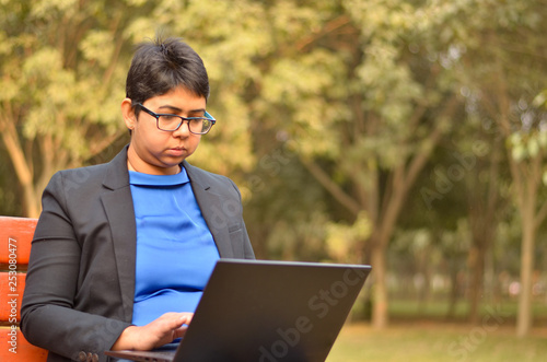Confident Indian professional business woman in business attire working on a laptop on a park bench © PhotographrIncognito