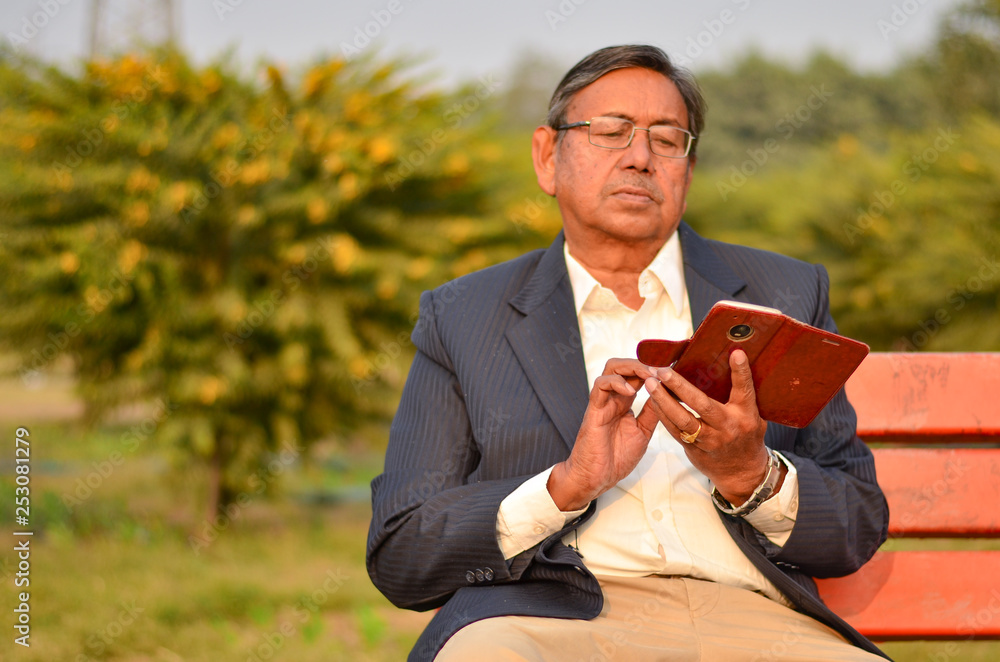 Senior Indian man curiously checking and working on his smart phone while sitting on a red bench in a park in Delhi, India Senior Indian man curiously checking and working on his smart phone while sitting on a red bench in a park in Delhi, India