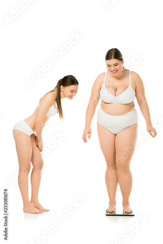 smiling slim woman in underwear looking at happy overweight woman on scales isolated on white, body positivity concept