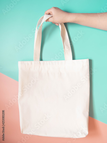 Blank white tote bag canvas fabric with handle mock up design. Close up of woman hand holding eco or reusable shopping bag on green orange background. No plastic bag and ecology concept. Copy space.