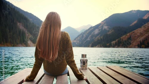 Long hair blond female traveler is doing mindful meditation sitting on bridge of lake green mountains in connection with nature Feel calm state of mind in relax moment of internal mental equilibrium