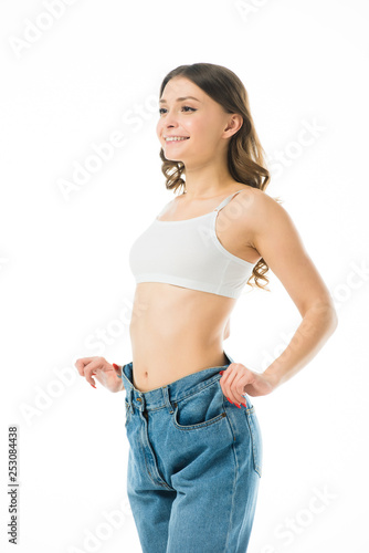 smiling slim young woman in big jeans isolated on white, lose weight concept © LIGHTFIELD STUDIOS