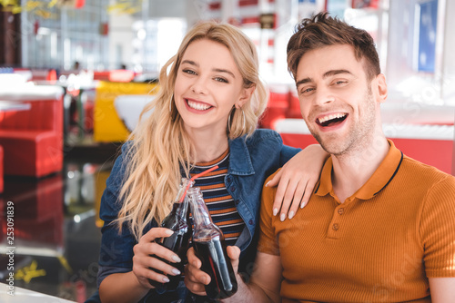 beautiful couple smiling, looking at camera and cheering with glass bottles at cafe