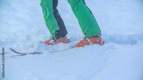 A man puts his skis on his feet and he waits on the snow. We can see his orange ski boots, too. photo