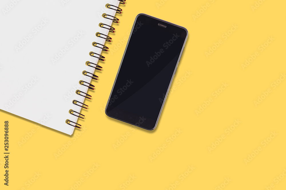 Black smartphone and blank paper notepad with spiral holder lies on yellow table in office or home. Top view. Business or education concept Black smartphone and blank paper notepad with spiral holder lies on yellow table in office or home. Top view. Business or education concept