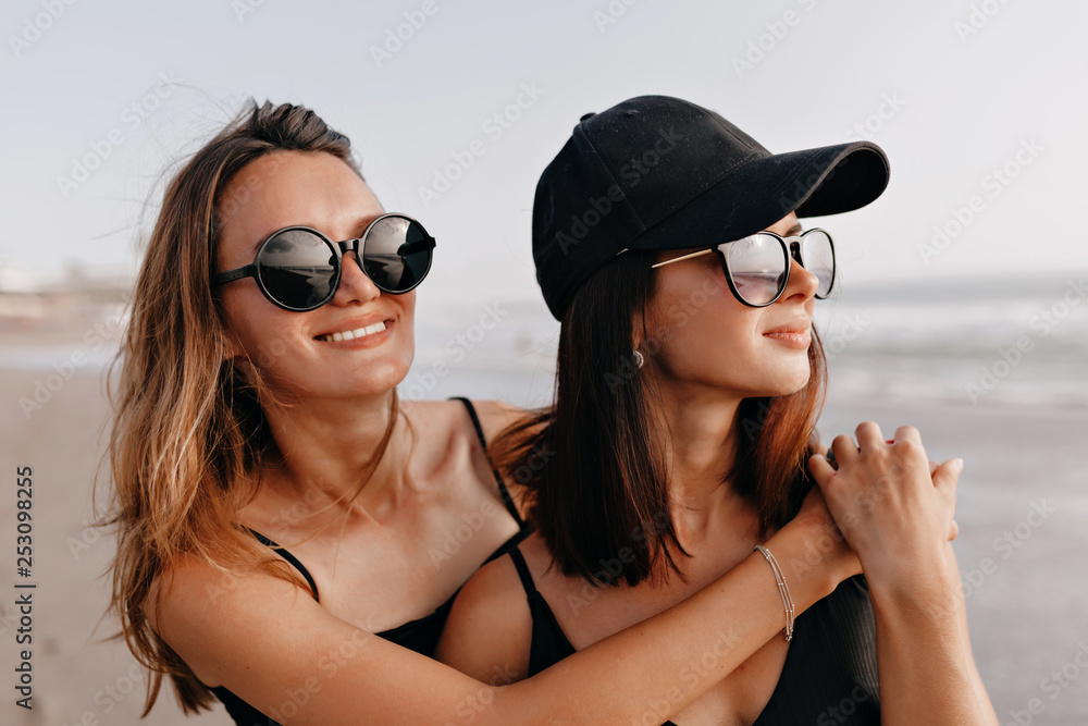 Pretty smiling young women walking on a beach. Two best friends strolling along the ocean shore on a summer day, enjoying vacation and looking dreamly on the ocean Pretty smiling young women walking on a beach. Two best friends strolling along the ocean shore on a summer day, enjoying vacation and looking dreamly on the ocean