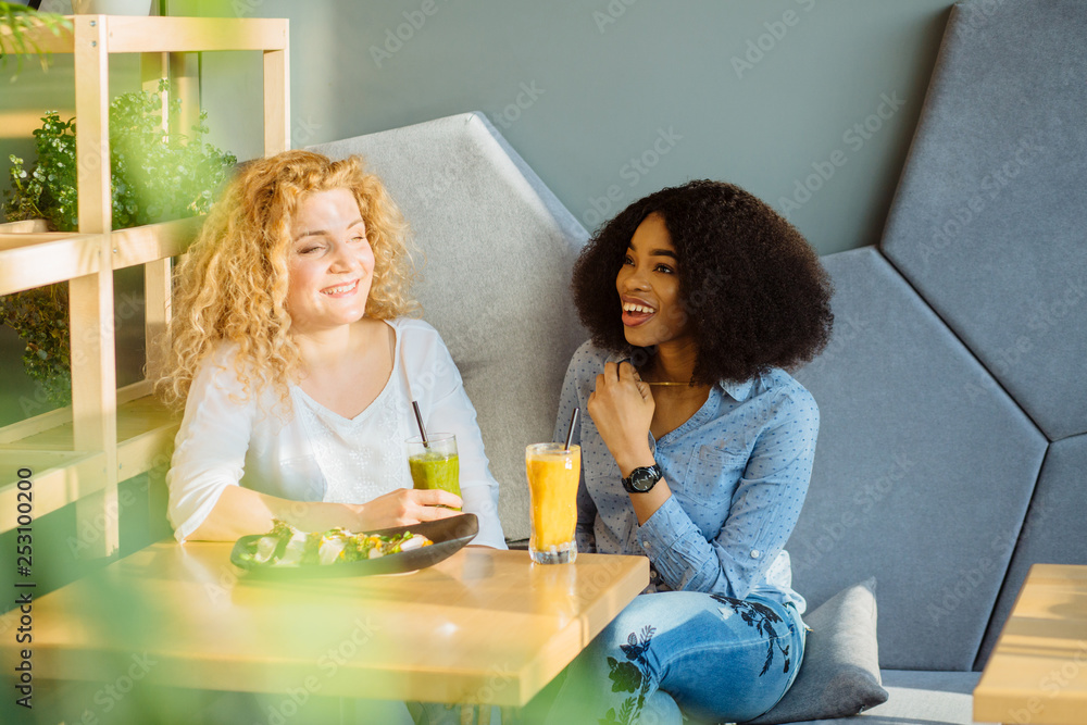 Two best friends enjoying nice conversation during lunch in modern cafe interior. African woman dining at restaurant together with white curly female. People, leisure, communication and friendship. Two best friends enjoying nice conversation during lunch in modern cafe interior. African woman dining at restaurant together with white curly female. People, leisure, communication and friendship.
