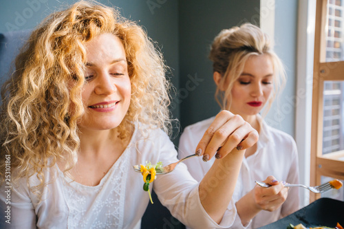 Blond curly woman eating fresh salad at restaurant. Healthy american girl with friend eating salad together looking away. Smiling young woman holding a forkful of salad. Health and diet concept.