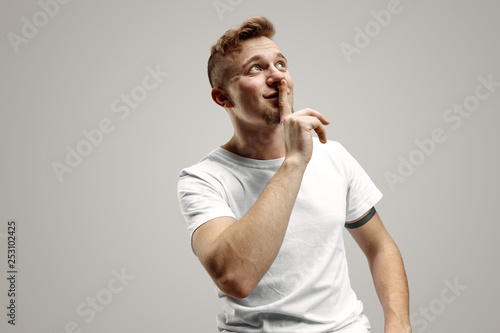 Keep silence. Handsome young man in white shirt looking at camera and holding finger on lips while standing against grey background