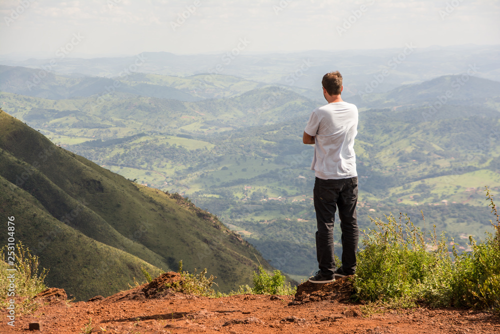 Young man seen from behind standing on the top of a mountain, admiring a green and mountainous landscape.
